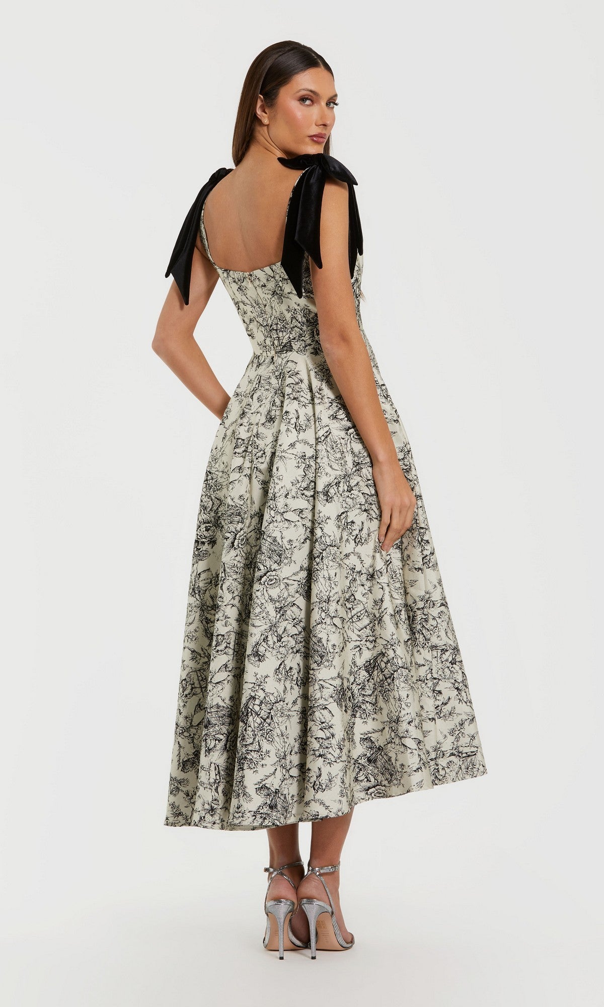A woman wearing the Mac Duggal 27749 Midi Party Dress—featuring floral brocade fabric and black ribbon shoulder straps—stands in heels, facing away from the camera against a plain white background, elegantly showcasing the sleeveless party dress.